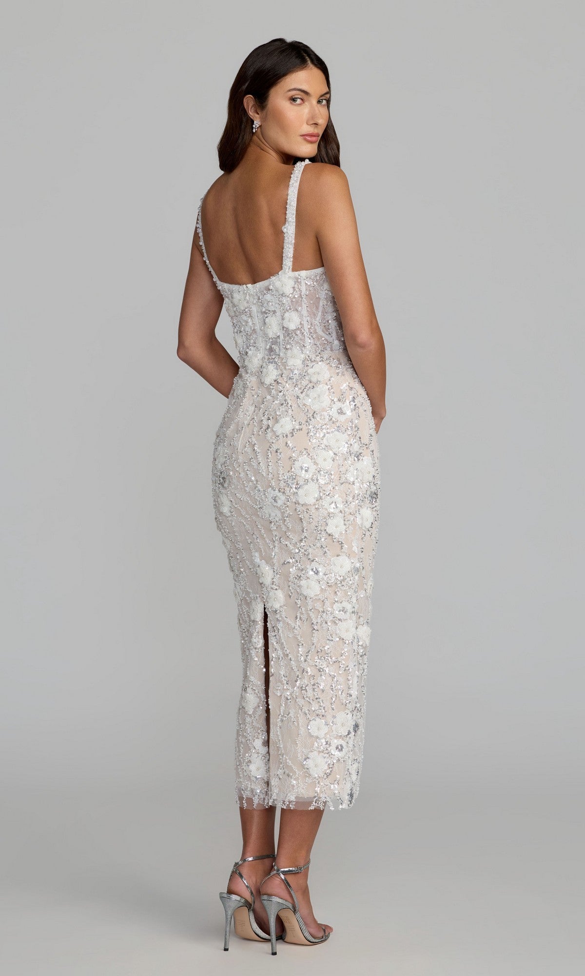 A woman stands facing away, looking over her shoulder in the Mac Duggal 21347 midi party dress—a sleeveless, sheer white lace dress adorned with delicate floral embellishments—paired with silver high-heeled sandals against a gray background.